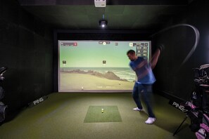 A person practicing golf in a room with a large screen displaying a golf course simulation, a green practice mat, and golf bags labeled 'PING'.