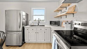 Une cuisine moderne dotée d’un réfrigérateur en acier inoxydable, de meubles blancs, d’une cuisinière noire et d’une fenêtre laissant entrer la lumière naturelle.