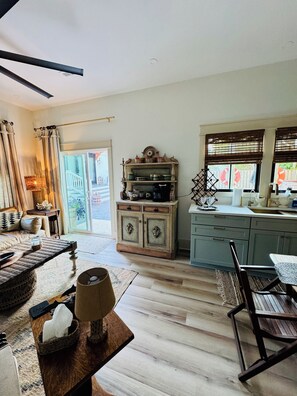 A spacious living area with a sofa, a coffee table, and a dining table. There is a kitchen area with a cabinet and a window with blinds.
