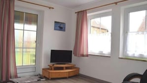 A room with a TV on a wooden stand, a door with curtains, and windows with curtains. - Ferienwohnung in Wenkendorf mit Garten (Fehmarn)