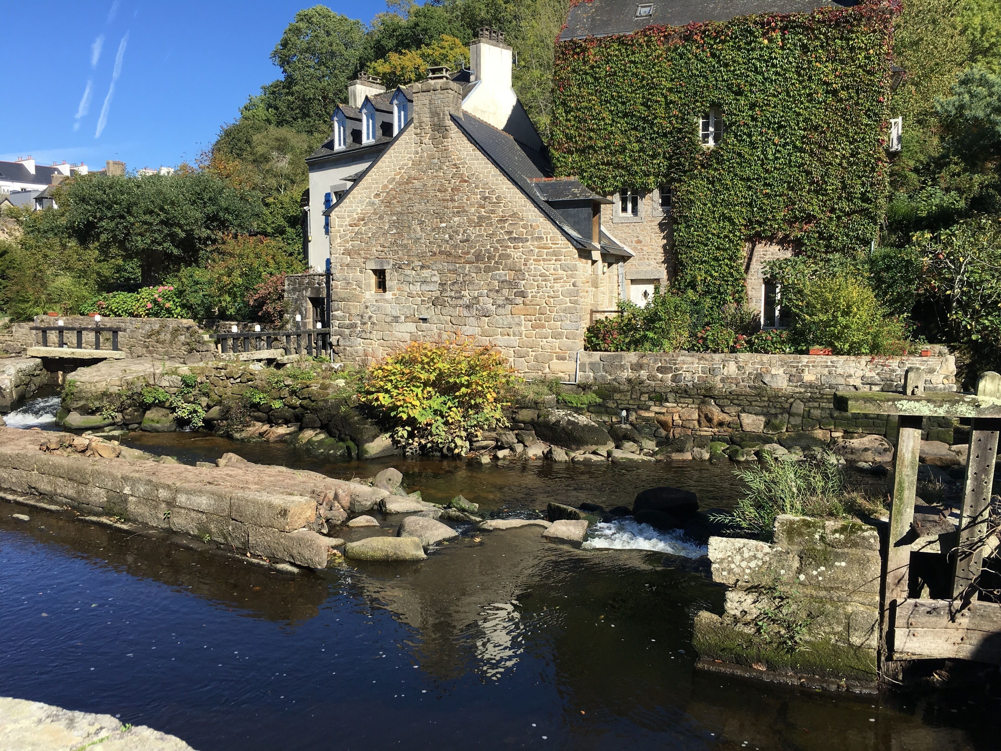 Un bâtiment en pierre avec un petit pont enjambant un canal d’eau, entouré de verdure et d’autres bâtiments.