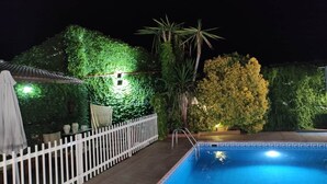 A swimming pool with a white fence, a covered seating area, and a wall of greenery at night.