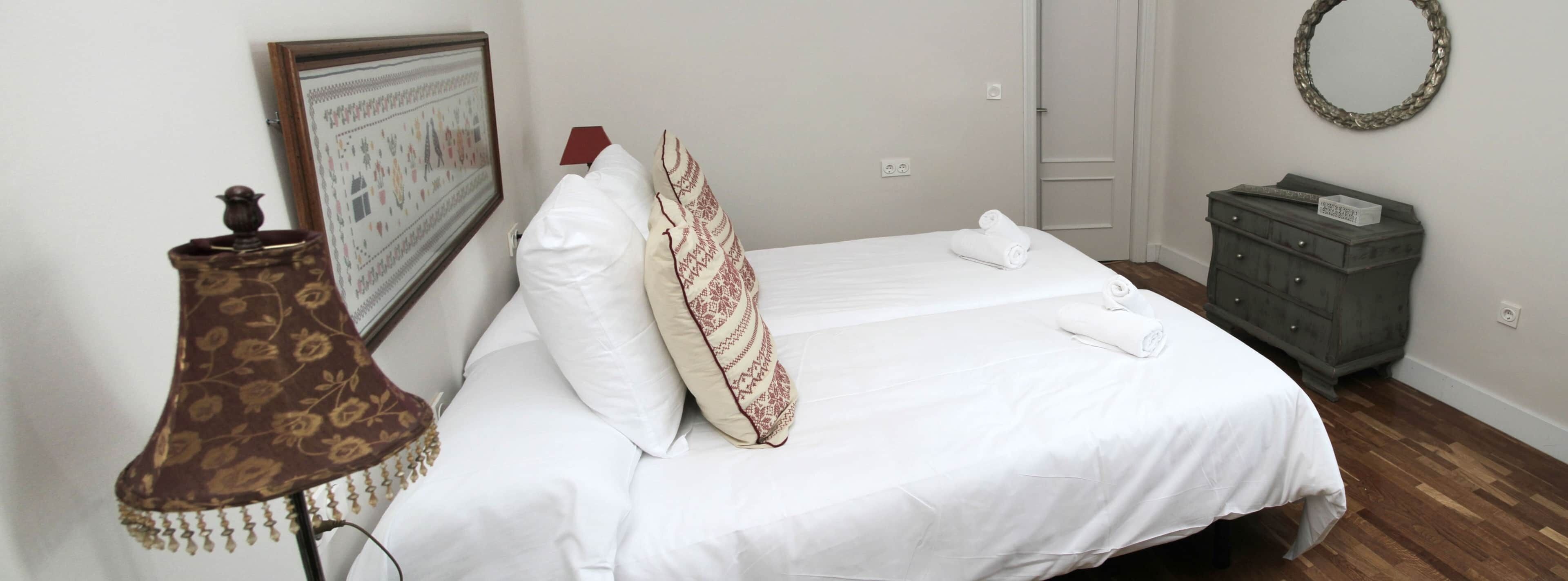 A neatly made bed with white linens, a patterned pillow, a small bedside table with a lamp, and a framed picture on the wall.
