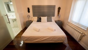 A neatly made bed with two rolled towels, a bedside table with a lamp, a nightstand with a lamp, a headboard, and a window with curtains. - Adapted room 1 in El Villar house (Estación de Brazatortas-Veredas)