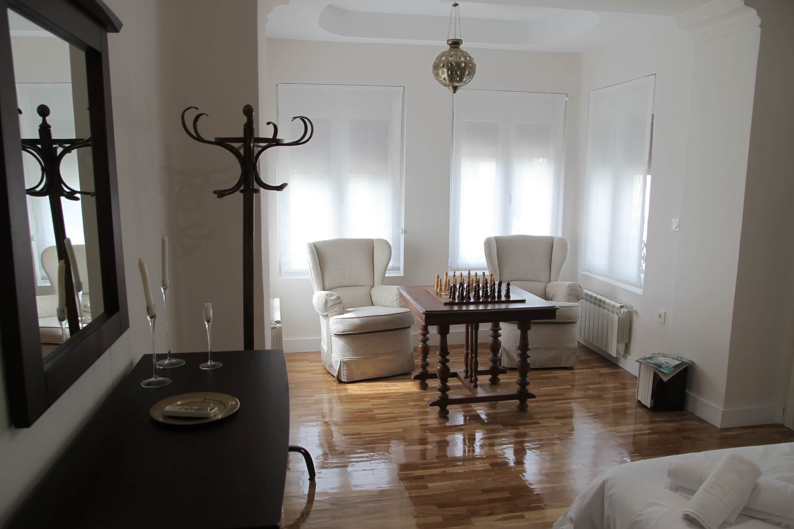 A room with a wooden floor, a chess table, two chairs, a mirror, and a chandelier.
