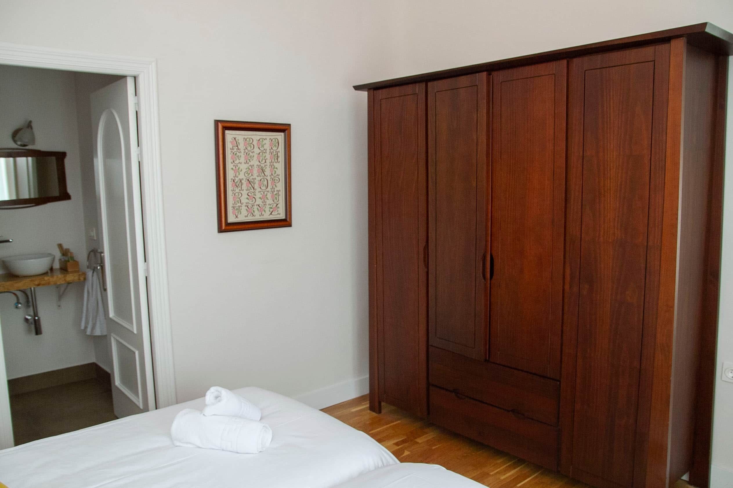 Ein Schlafzimmer mit Holzschrank, einem Bett mit weißer Bettwäsche, einem gerahmten Kunstwerk mit Alphabet und einem Badezimmer mit Waschbecken und Handtuchhalter.