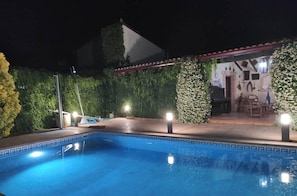 A swimming pool with a geometric tile pattern, surrounded by greenery and a covered patio area with seating.