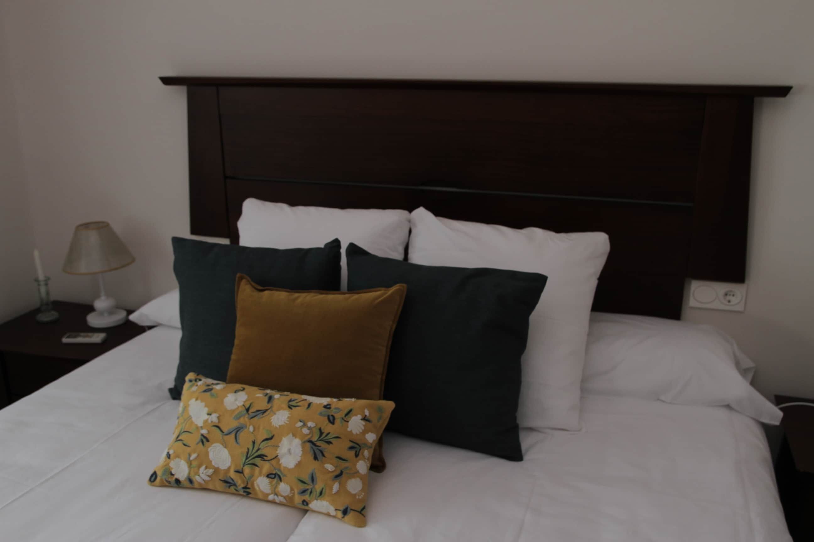 Una cama con ropa de cama blanca y varios almohadones, una mesita de noche con lámpara y un cabecero de madera oscura.