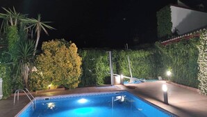 A swimming pool with a tiled edge, surrounded by greenery and a white building in the background.