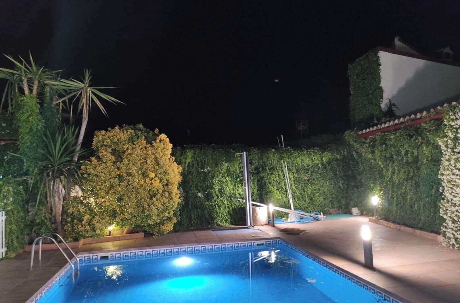 Una piscina con borde de azulejos, rodeada de follaje y un edificio blanco al fondo.