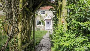 A stone pathway leading to a white building with a red roof, surrounded by lush greenery and a wooden arbor.
