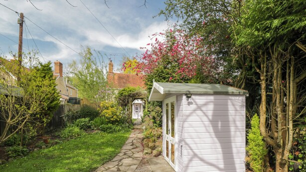 A stone pathway leading to a white shed in a garden with various plants and a brick house in the background. - Lavender Cottage (Gillingham)