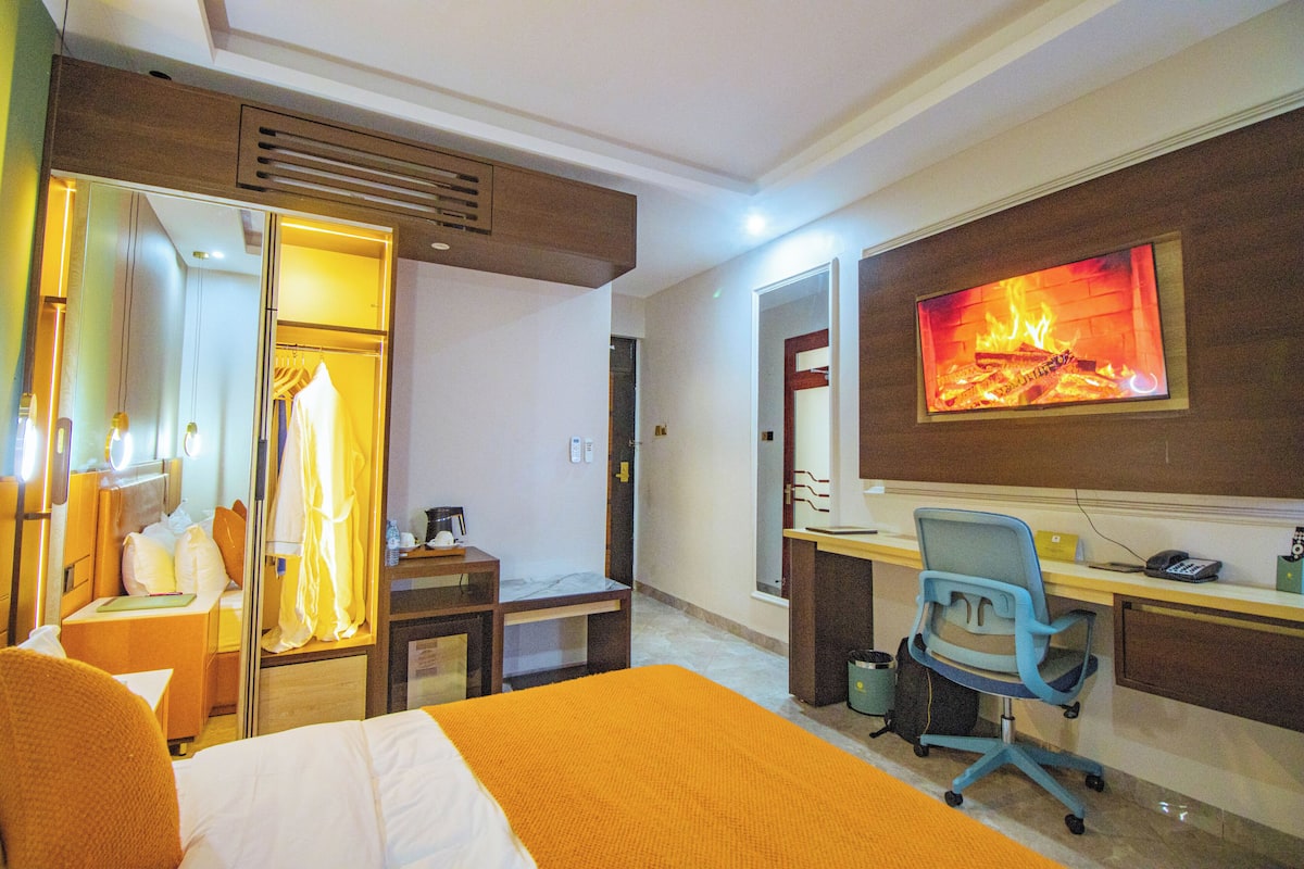 Habitación de hotel con cama, escritorio con silla, armario y televisión mostrando una chimenea.