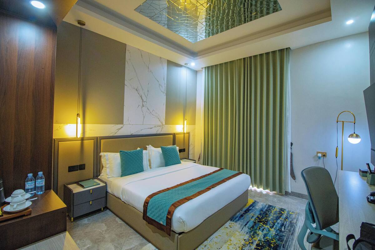 Habitación de hotel con cama, mesitas de noche, escritorio, silla, lámpara de escritorio y ventana con cortinas.