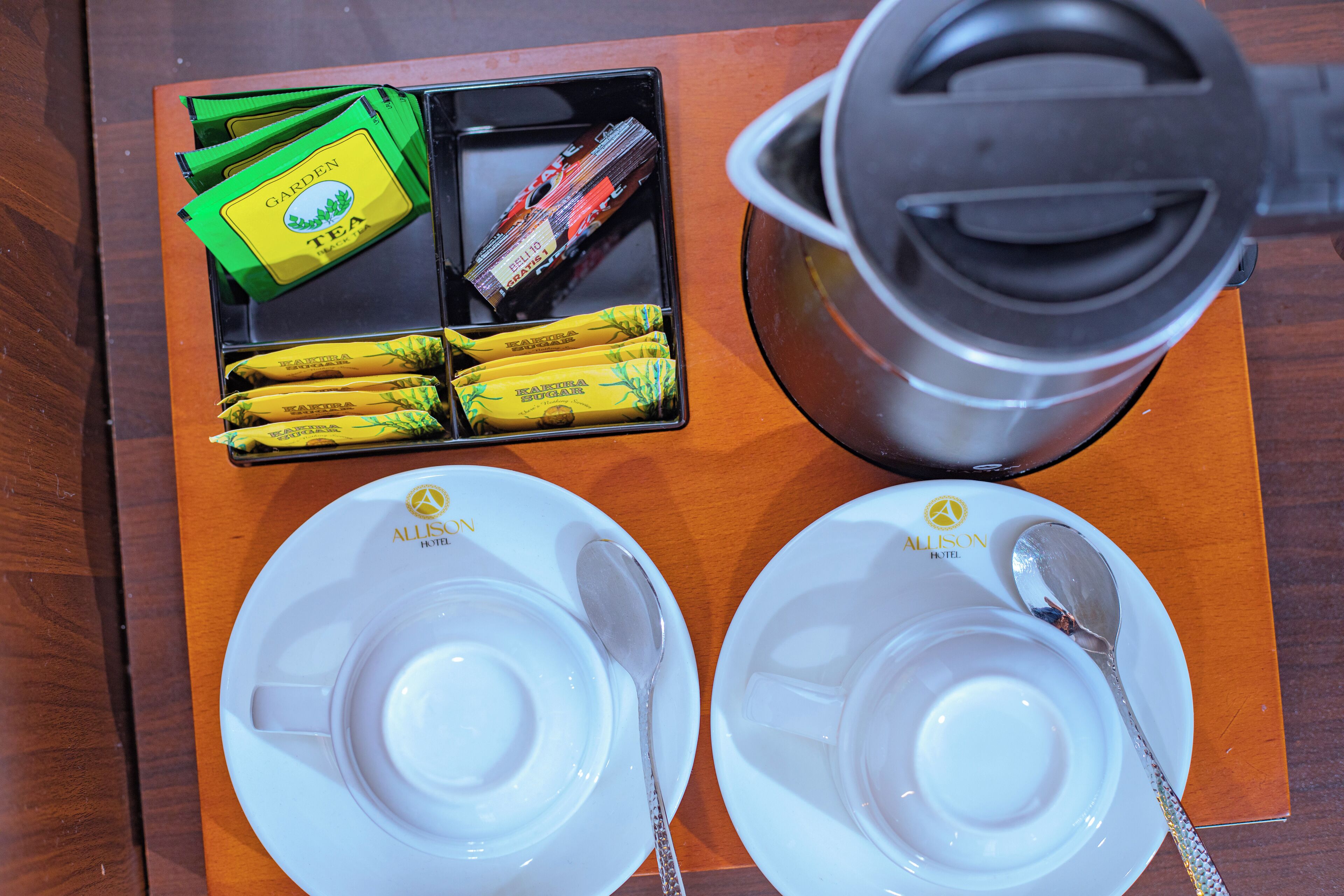 Un plateau avec des sachets de thé et une bouilloire, deux assiettes blanches vides accompagnées d’une cuillère, et le logo de l’hôtel.