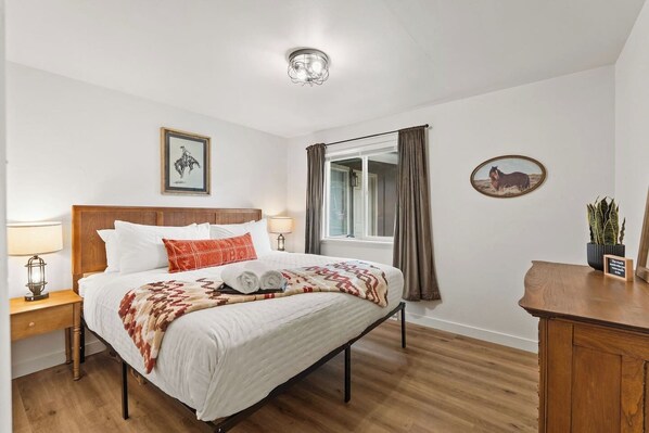 Une chambre à coucher bien rangée, avec un lit en bois, deux lampes de chevet, une fenêtre avec des rideaux et un tableau accroché au mur.