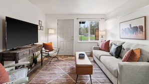 Una sala de estar con una televisión, un sofá, una mesa de centro, un sillón y una puerta que da a un balcón.