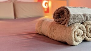Ein ordentlich bezogenes Bett mit einem aufgerollten, beige gefärbten Handtuch, eine rosa Wand und ein weißer Schrank im Hintergrund.