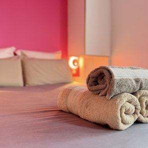 Un lit bien fait, avec une serviette beige enroulée posée dessus, un mur rose et une armoire blanche en arrière-plan.