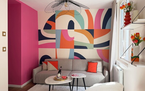 Un salon moderne agrémenté d’une œuvre d’art abstraite aux couleurs vives, d’un canapé gris avec des coussins orange, d’une petite table basse ronde et d’une coupe de champagne.
