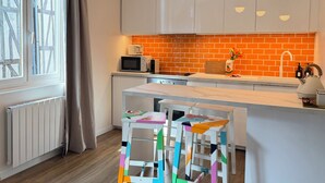Eine moderne Küche mit weißen Schränken, einer Ziegelrückwand, einer Mikrowelle, einem Toaster und einem Spülbecken. Bunte Hocker und ein Fenster mit Vorhängen sind ebenfalls vorhanden.