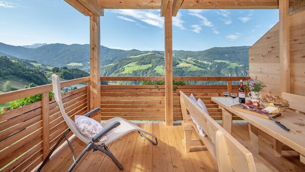 Um deck de madeira com vista para colinas verdes, um banco, uma mesa com comida e bebidas, e uma cadeira.