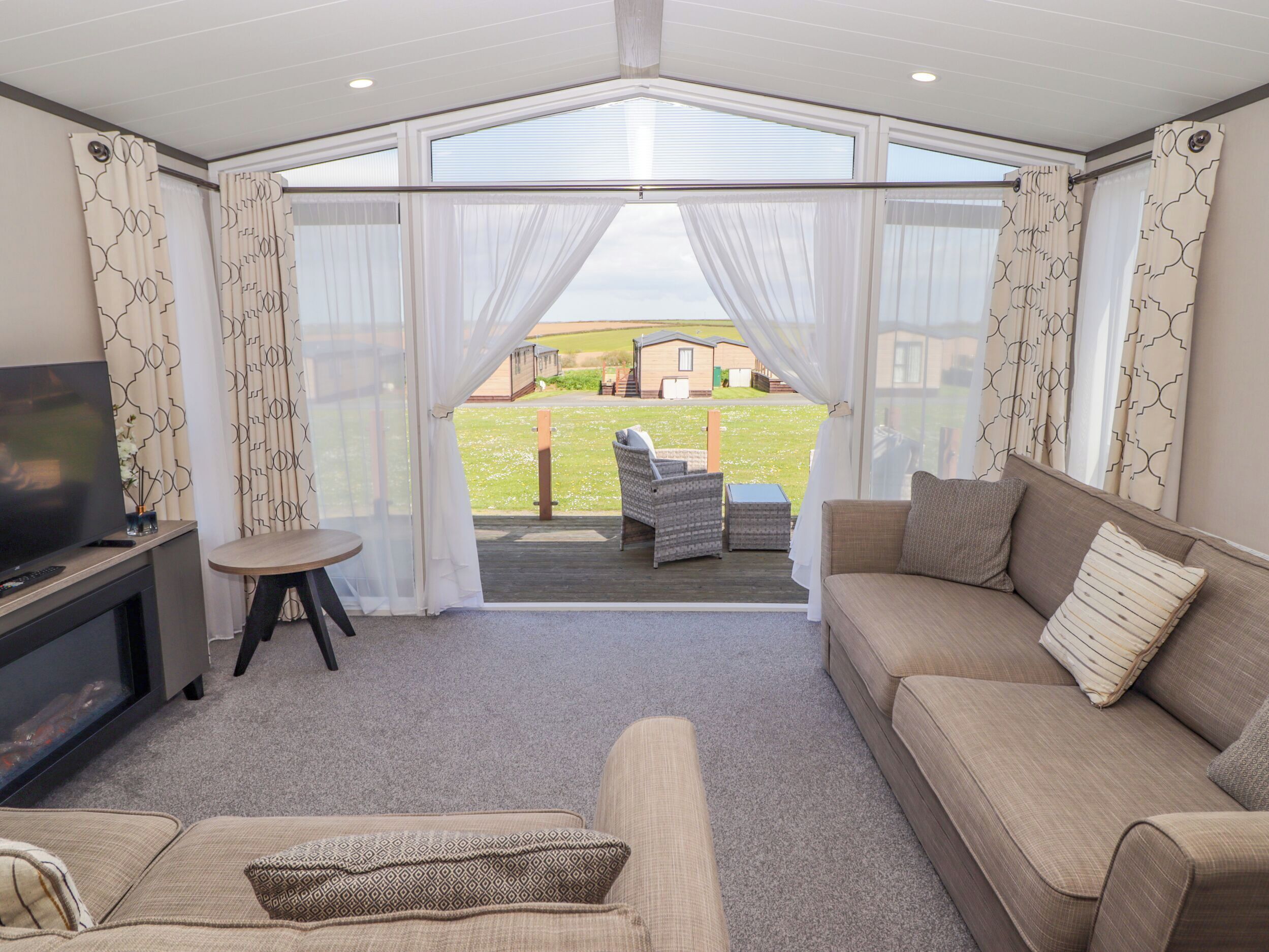 A cozy living room with a TV, sofa, and a small table. There is a balcony with a view of the countryside.