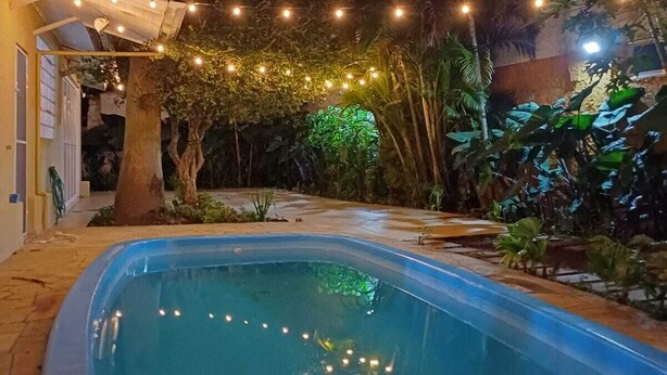 En swimmingpool med lyskæder, omgivet af tropiske planter og et overdækket område.