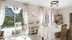 A bright room with a dining table, chairs, a white console table with decorative items, and a view of a garden through large windows. - Rêves d'Emeraude – Le Rêve du Pêcheur Guest Room (Cancale)