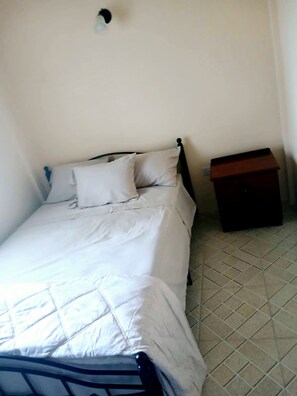 Una cama individual con estructura metálica, ropa de cama blanca y una pequeña mesita de noche a su lado.