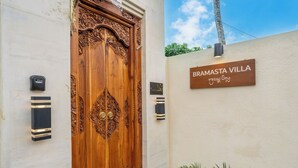 Una puerta de madera con intrincados grabados, un cartel que dice 'BRAMASTA VILLA' y dos lámparas fijadas a la pared.