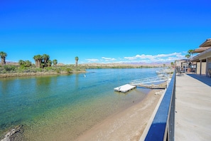 A clear river with a dock, palm trees, and a building with a balcony. - Riverfront w/ Dock & Desert Views 4BR + Media Room, BBQ & Garage (Bullhead City)