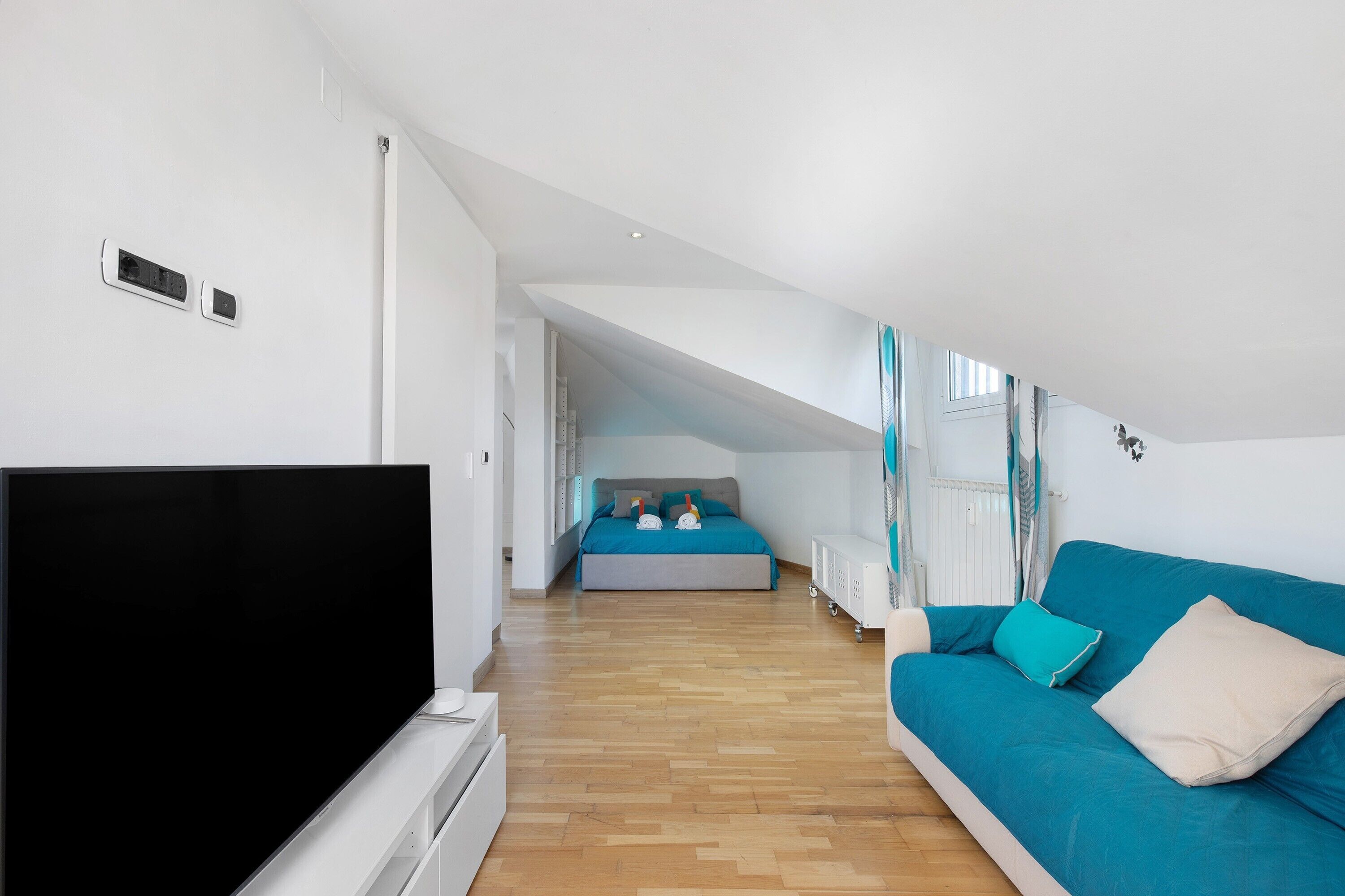 A modern, well-lit room with a flat-screen TV, a blue sofa, and a bed with teal bedding.