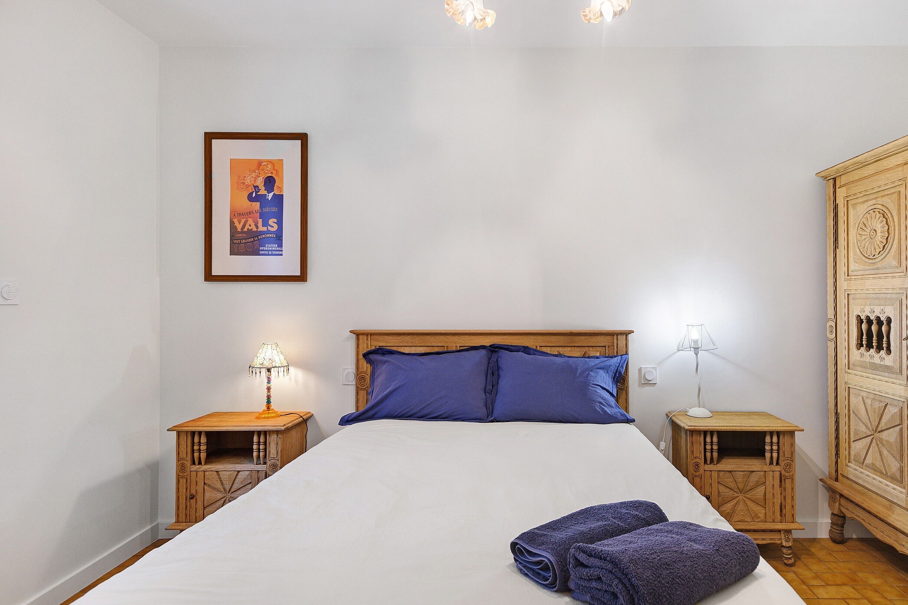 A neatly made bed with a white sheet, two blue pillows, and a folded towel on the right side. There are two wooden nightstands with lamps, and a wooden wardrobe with a decorative panel on the right.