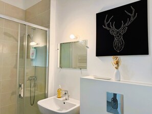 Une salle de bain avec une douche en verre, un lavabo blanc, un miroir et une œuvre d’art représentant une tête de cerf, accrochée au mur.