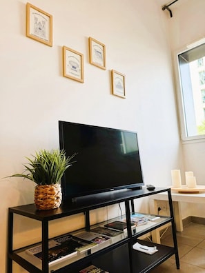 Une pièce avec un téléviseur à écran plat posé sur un meuble noir, une petite plante, des certificats encadrés accrochés au mur et une fenêtre donnant sur un paysage.