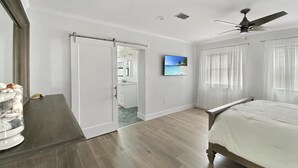 A bedroom with a bed, a ceiling fan, a TV, and a bathroom visible through an open door. - Saltwater Pool & Spa Villa on a Gulf-Access Canal (Englewood)