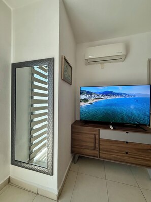 Ein Zimmer mit einem großen Flachbildfernseher auf einer Holzkommode, einer Wandklimaanlage und einem großen Spiegel mit geometrischem Muster.