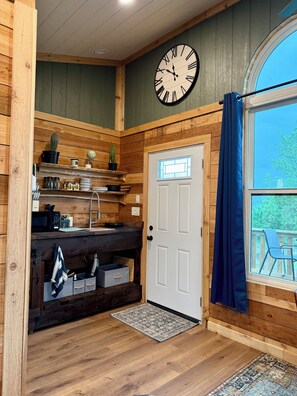 A rustic kitchen with wooden walls, a clock, a door, and a window with a view. - Unique 10-Acre Escape: Treehouse Studio, Fishing Ponds, Fire Pit, Pizza Oven (Grain Valley)