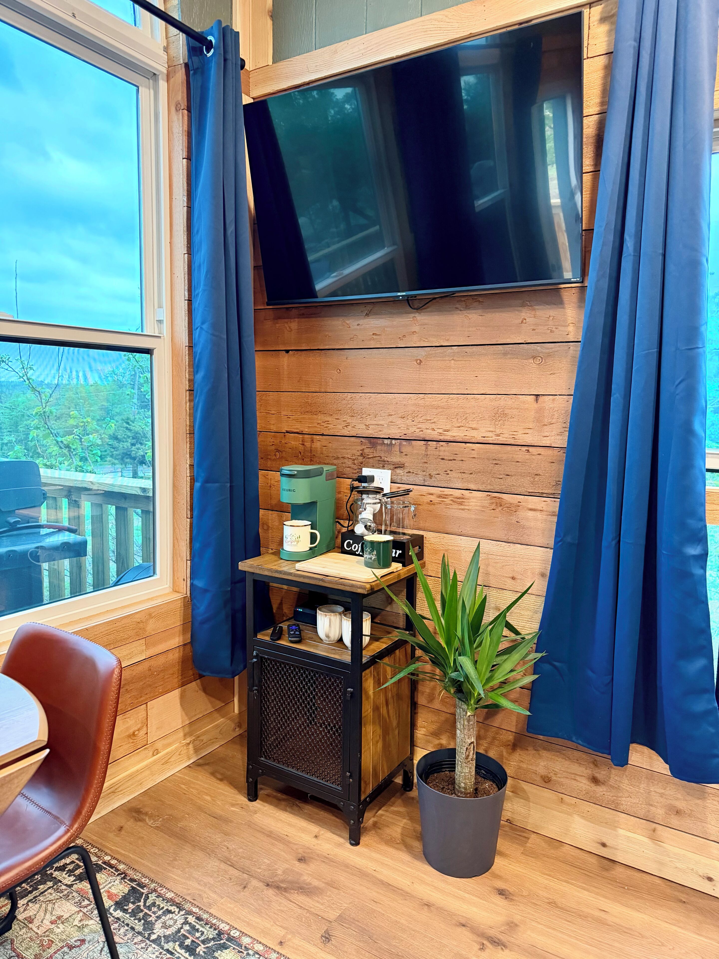 A cozy room with a wooden wall, a TV mounted above a table with a coffee maker, a potted plant, and a chair.