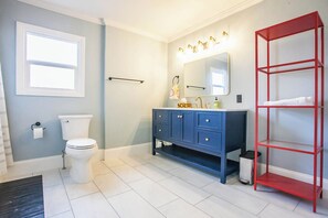 A bathroom with a toilet, a blue vanity with a mirror and lights, and a red shelf. - Junebug - 2 bedroom, 1 bathroom home in The Heights (Hood River)