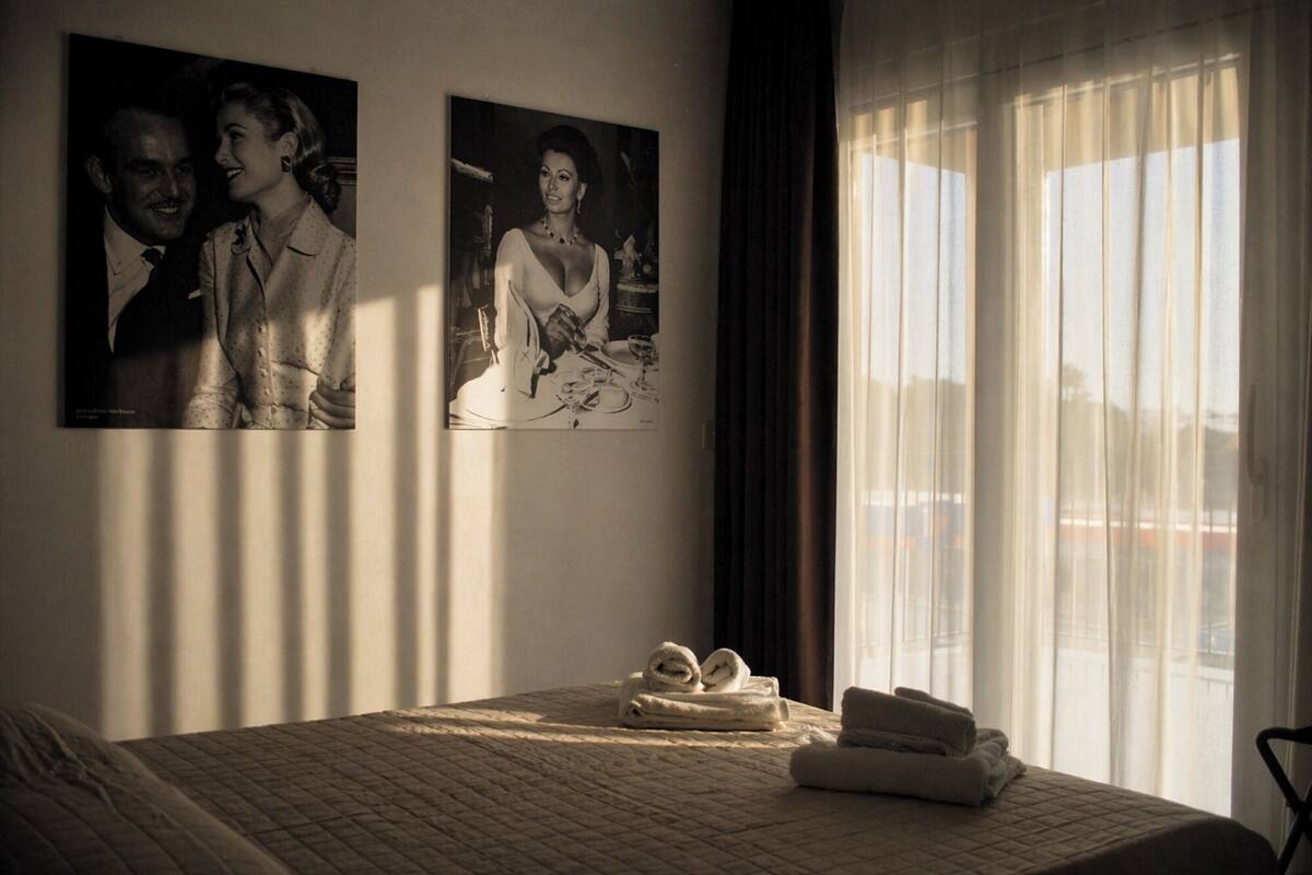 A hotel room with a bed, two framed pictures on the wall, a window with curtains, and a towel on the bed.