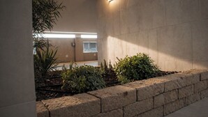 Un pasillo con pared de piedra, una jardinera de ladrillo bajo con plantas y una lámpara fijada a la pared.