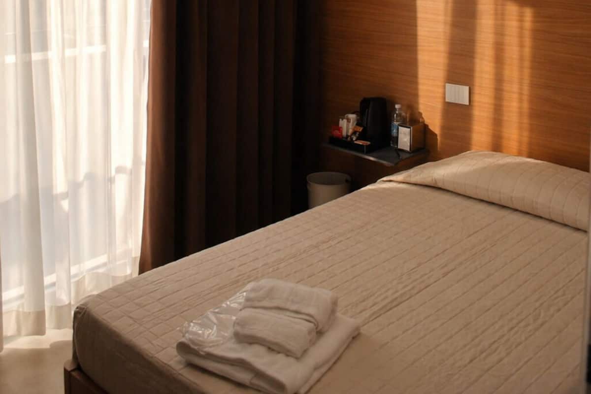 A hotel room with a bed, a nightstand, a wooden headboard, a window with sheer curtains, and a modern ceiling light fixture.