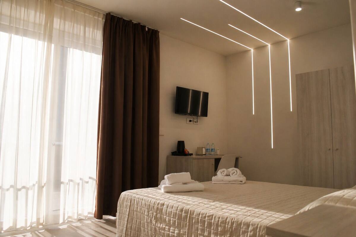 A hotel room with a bed, a TV mounted on the wall, a desk with a chair, and a wardrobe.