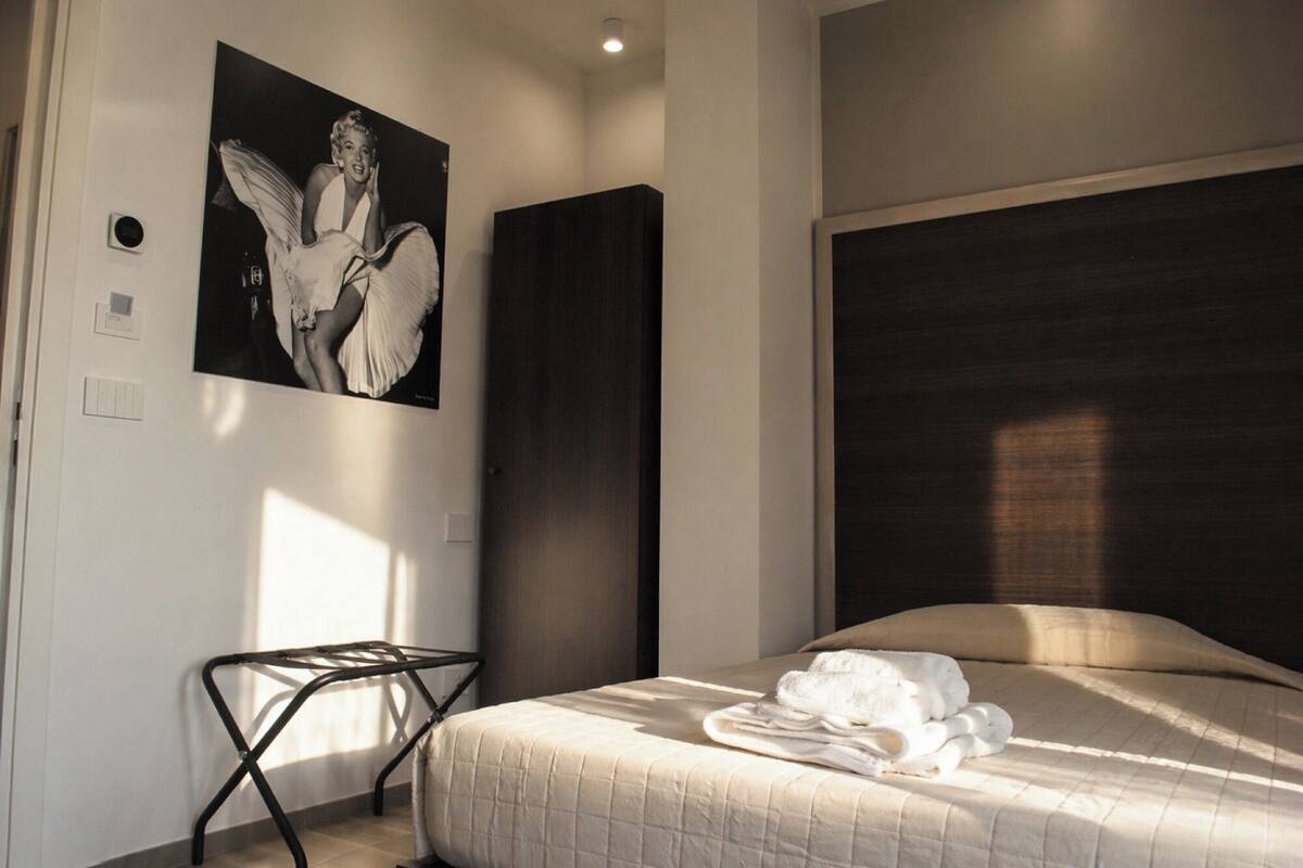 A bedroom with a bed, a black metal chair, a large dark headboard, and a framed picture on the wall.