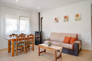 A living room with a sofa, a dining table with chairs, and a small table with a plant. There are three wall-mounted pictures and a black heater.