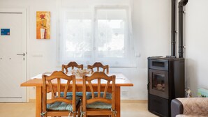 Ruokailutila, jossa on puinen pöytä ja tuolit, ikkuna, jossa on läpikuultavat verhot, musta liesi ja ovi, jossa on kyltti.