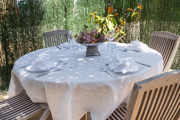 A round table with a white tablecloth, surrounded by wooden chairs, set for a meal with cutlery and a vase of flowers.