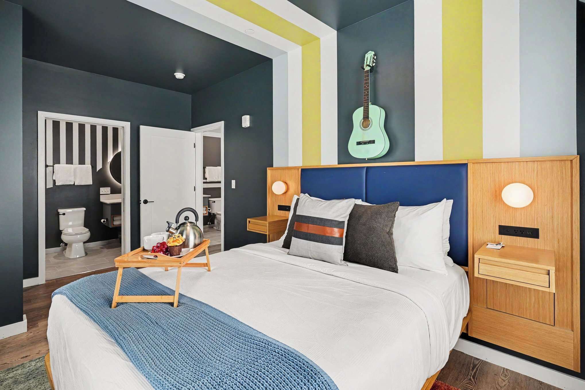 A modern hotel room with a large bed, a wooden headboard, a bedside table with a lamp, and a guitar on the wall.